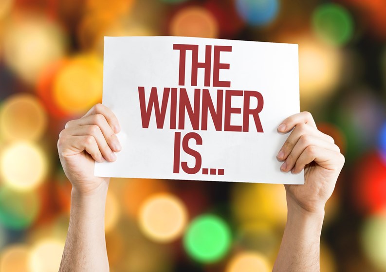 Two hands holding a sign reading THE WINNER IS...in front of a blurred multicolored background
