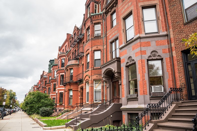 Row of old red-brick apartment buildings with staircases to the front doors along a tree lined street on a cloudy autumn day. Boston, MA, USA.